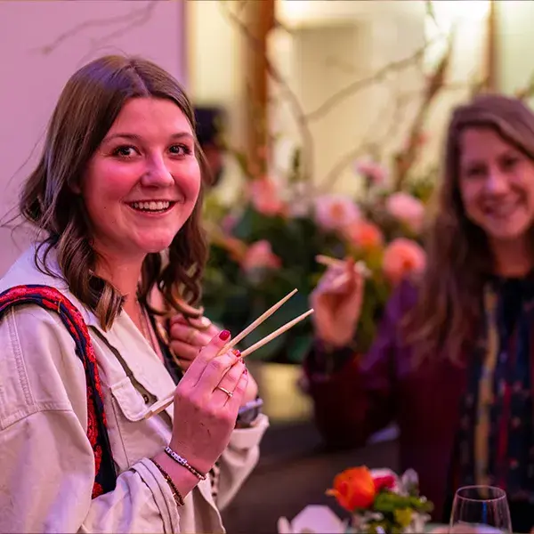 A picture of two smiling female guests at a small party, showing off their chopstick skills
